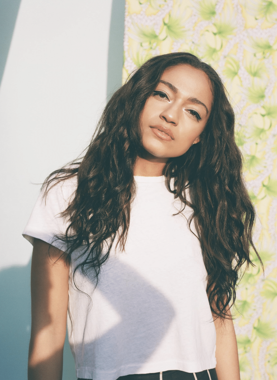 A woman with long, wavy dark hair wearing a white t-shirt stands in front of a colorful, patterned backdrop, looking at the camera with a relaxed expression—a scene straight from this season’s Pop Culture Picks. Sunlight casts soft shadows on her and the wall.